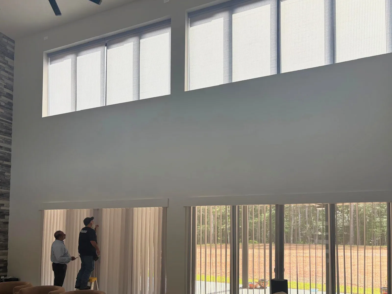 Two installers at the patio doors of a two-story great room, one on a step stool at the vertical blinds, with shades already fitted to the windows high above.