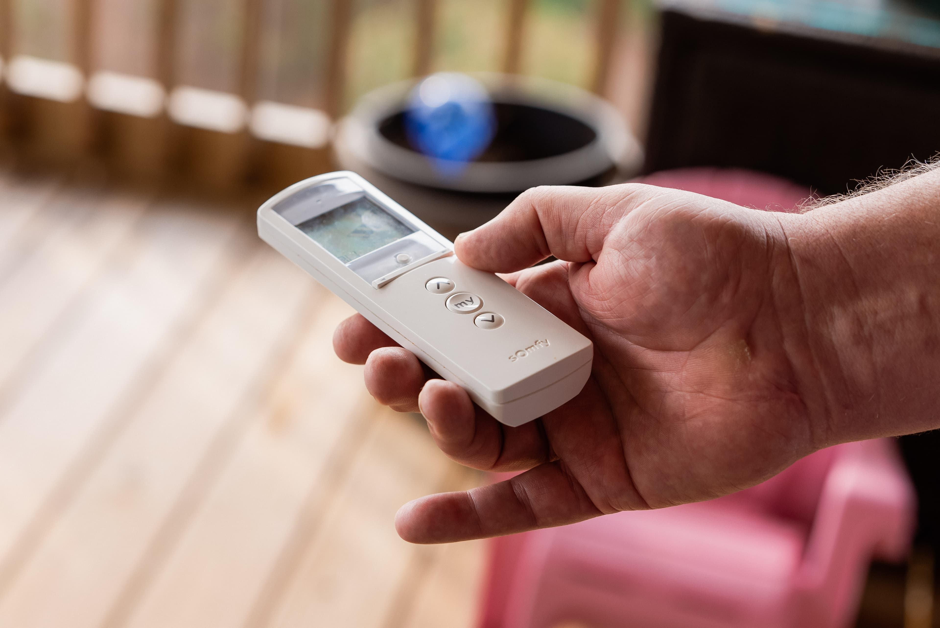 A hand holding a white five-button remote control for motorised shades, thumb resting on the up button.