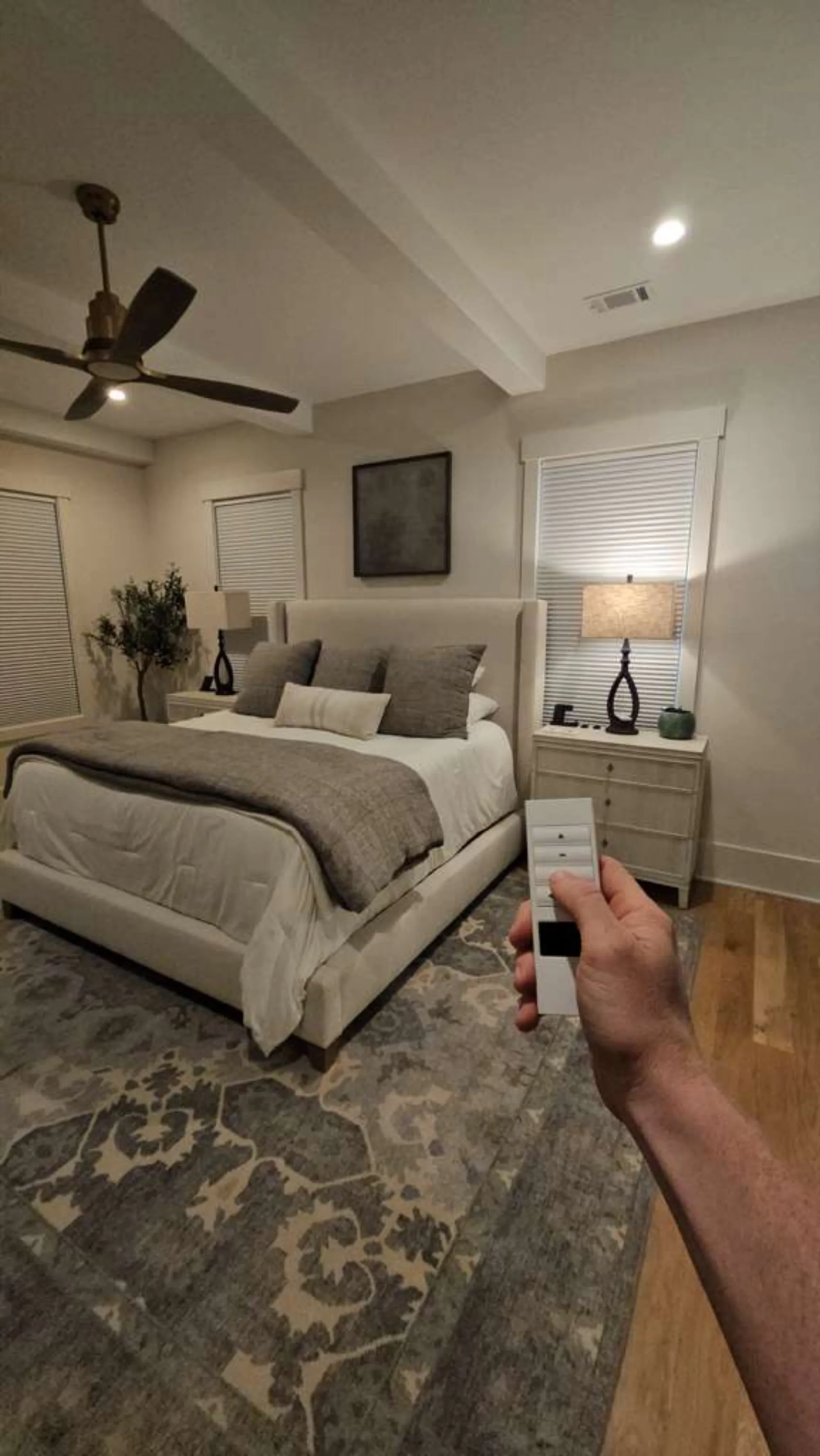 A hand holding a shade remote in a bedroom, with white cellular shades lowered on the windows.
