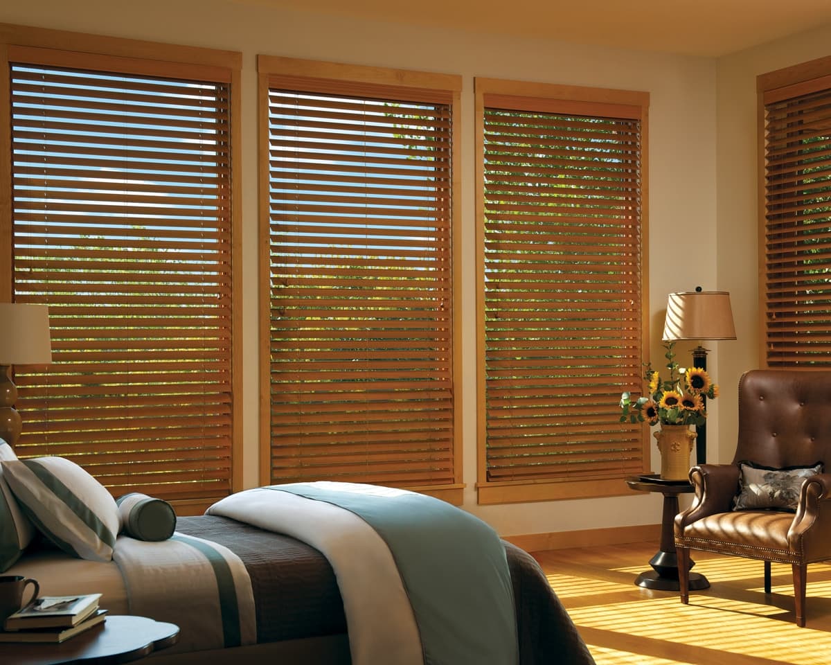Wood-grain blinds on a row of tall windows in a sunlit bedroom.