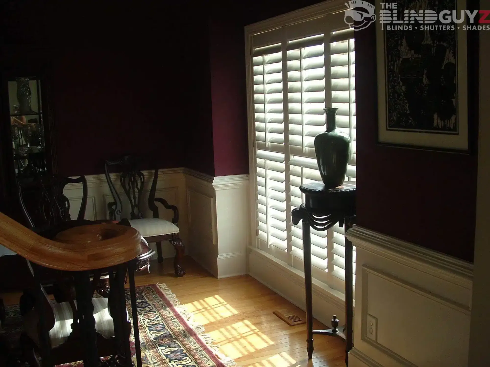 White plantation shutters on a tall window in a dining room with deep red walls and wainscoting.