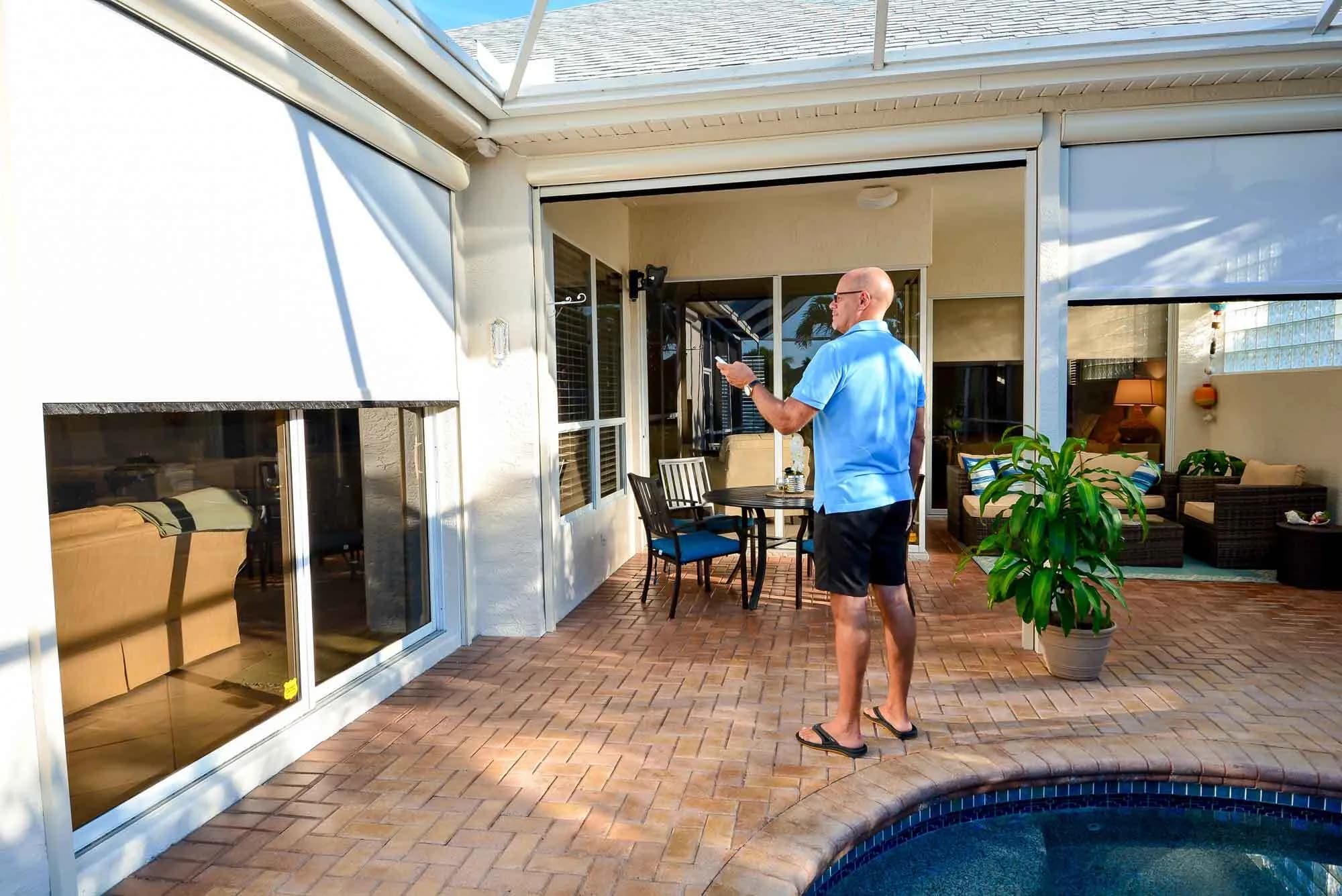 A man on a poolside patio pointing a remote at a retractable screen that is part-way down across a wide opening, with a second screen already closed beside it.