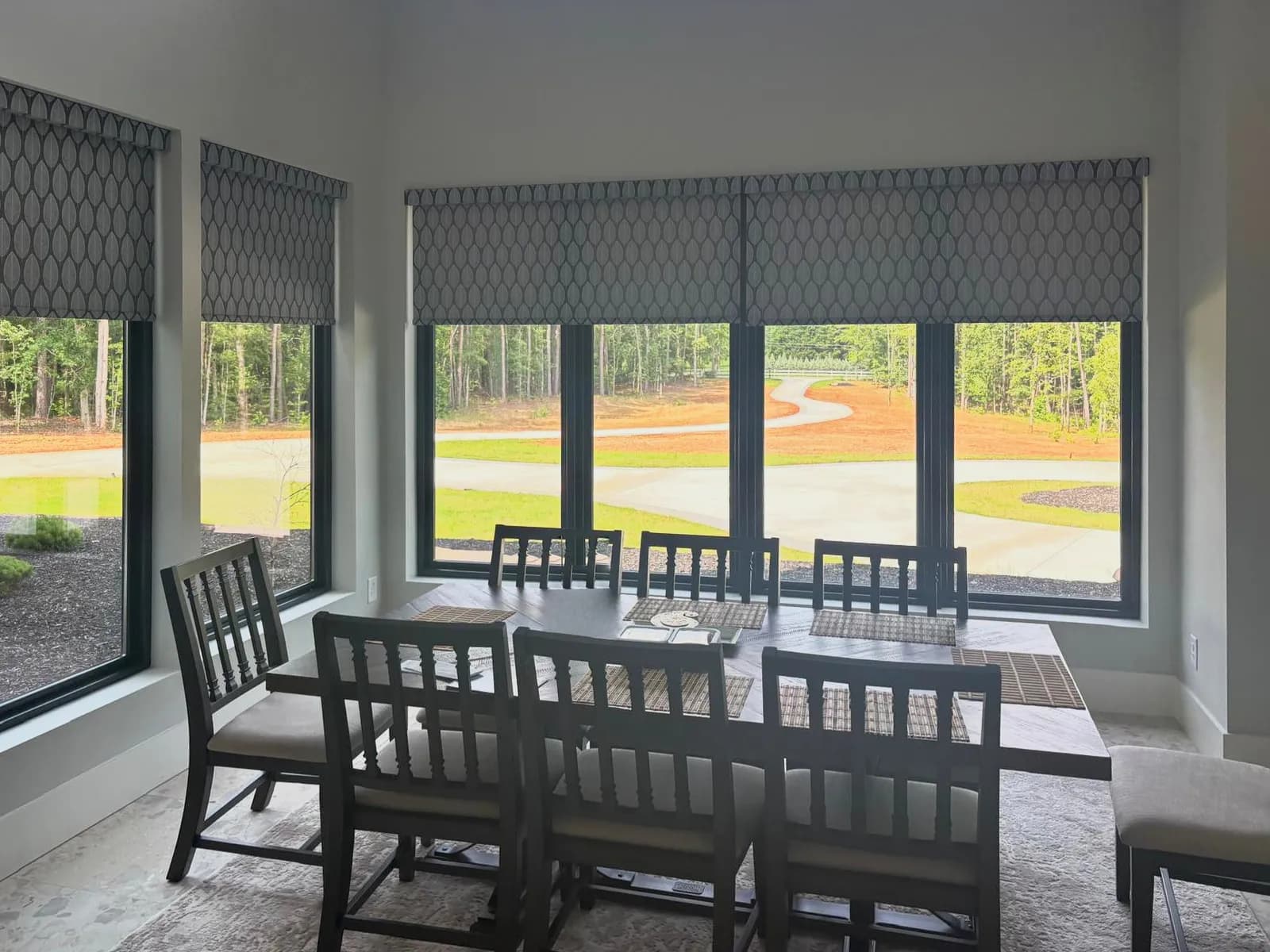 Grey roller shades with a white leaf pattern, lowered partway on the corner windows of a dining room that looks out over woods.