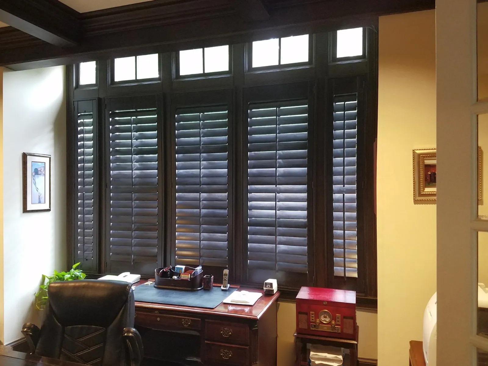 Dark stained wood plantation shutters across a bank of windows in a home office, with the transom windows above left bare.