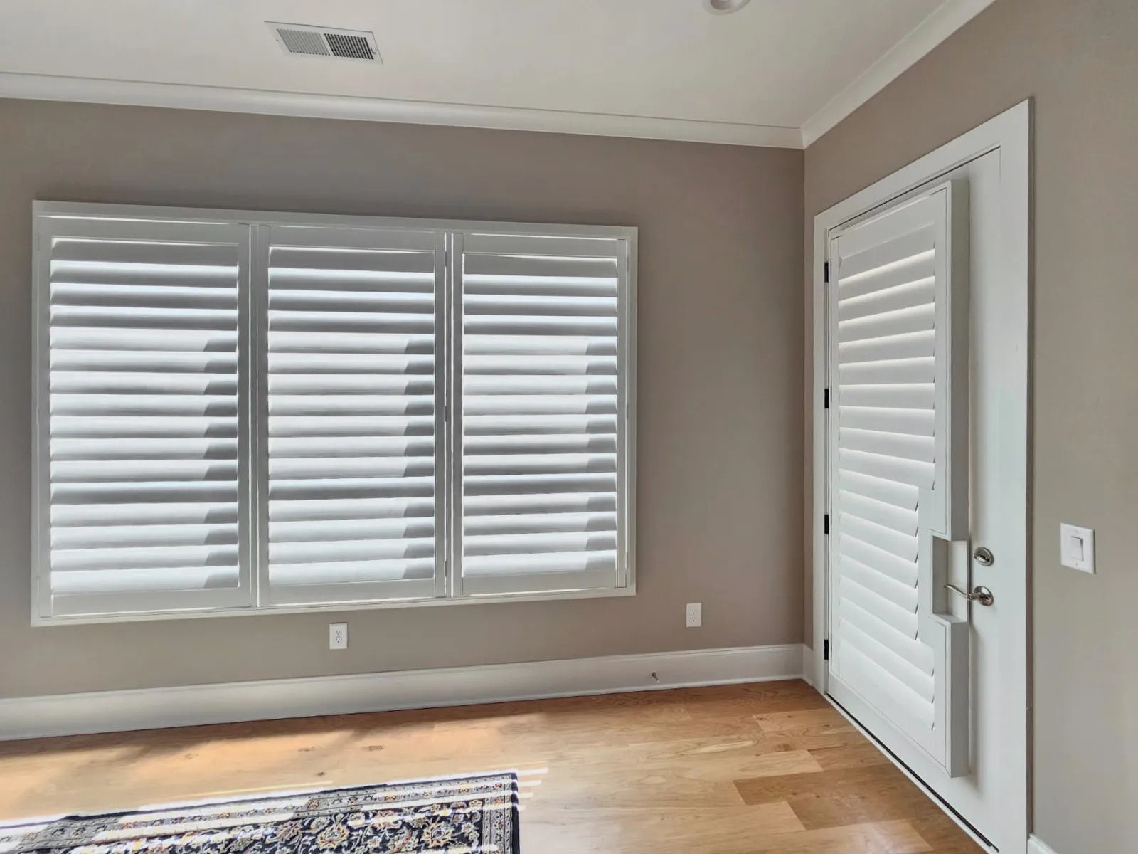 White plantation shutters on a triple window and on the door beside it, the door's panel cut out around the handle.